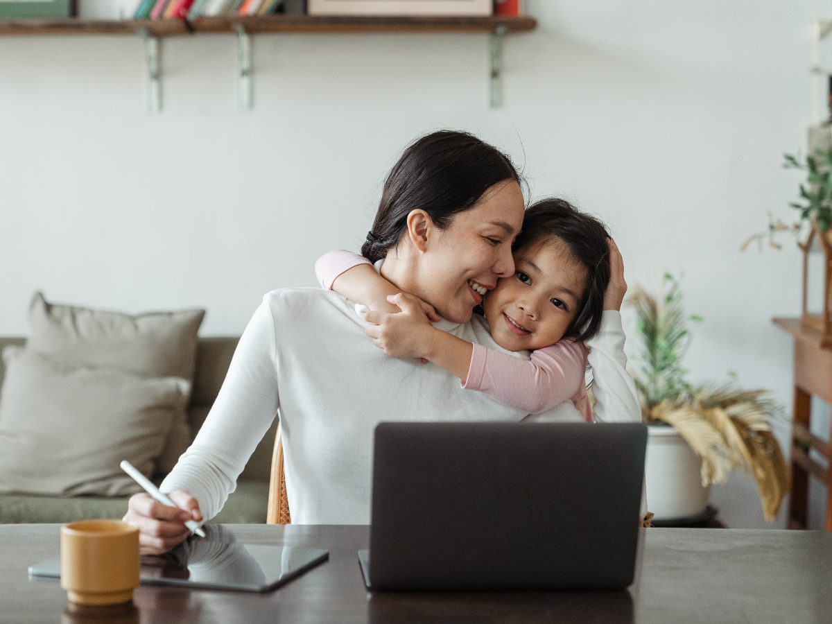 woman sitting at desk with computer open, smiling and hugging her daughter